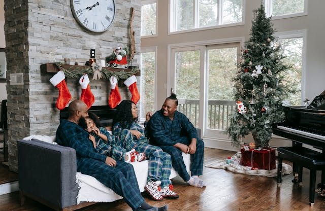 a family sitting on the couch n front of a fireplace with stockings and a christmas tree.